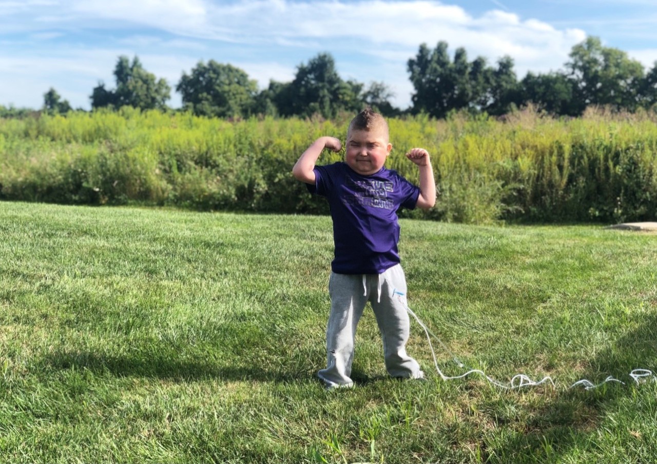 Image of Cohen in a purple shirt standing outside in backyard, flexing his muscles like a superhero